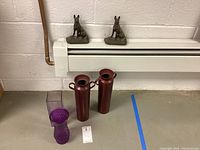 Two cast iron dog door stops on a white baseboard, two metallic burgundy vases, and two purple glass vases on the floor in front