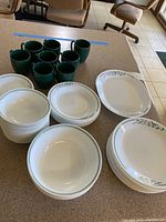 Corelle dishware stacked and arranged showing plates, bowls, and green mugs with floral rim design