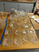 View of assorted crystal and etched glass serving dishes, vases, pitcher, ice bucket, and trays arranged on a wooden table.