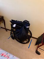 Folded Drive Cruiser III wheelchair positioned by the wall on carpeted floor, showing size and folded form with visible rear wheels and part of the armrest.