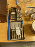 Top view of boxed sets of knives, forks, wooden spoon and fork alongside cups and saucers on table.