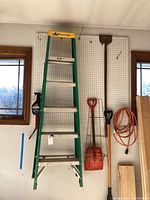 Full view of green Louisville 6ft utility ladder and surrounding yard tools hanging on pegboard.