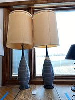 Pair of tall ceramic lamps with fabric beige drum shades in front of window. Both lamps have grey ceramic bases with embossed oval pattern.