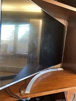 Side view showing curved silver stand and black screen with reflections, the TV is large and placed on wooden furniture.