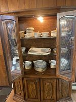 Full view of wooden china cabinet with glass doors open showing stacked Mikasa china pieces inside, including plates and bowls.