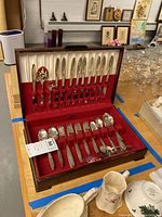 Closed wooden case holding Oneida Community silver plated flatware set on red velvet lining. Includes knives and serving utensils.