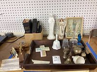 Photo showing religious books including bibles on left, several figurines including a tall white Mother Mary statue, two angel figurines, a framed print, various bells in the wooden tray, and a ceramic blessing cross.
