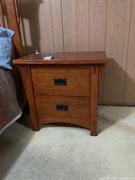 Front view shows wooden nightstand with two drawers and black handles, placed on carpet next to bed and wall with vertical paneling.