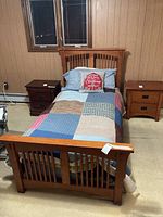 Solid wood single panel bed frame dressed with patchwork quilt and Christmas pillow, flanked by two wooden nightstands, one on each side. Room has wood-paneled walls and beige carpet.