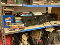 Wide shot of metal shelving with Sony CD player, RCA receiver, and multiple VHS/DVD media on shelves.
