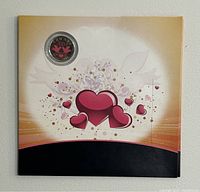 Front view of the decorative card packaging showing the coin mounted in the top left corner with heart and floral designs.