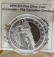Front side of 2014 $10 silver coin showing a cowboy riding a horse with mountains in background, in protective capsule.