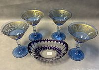 Four blue stemmed wine glasses with gold detailed floral and scroll patterns around bowls along with a purple cut glass thumbprint oval bowl on a neutral surface.