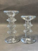 Front view of two clear textured glass candleholders showing the ringed segmented pattern, one taller with two rings and one shorter with three rings, set on a neutral background.