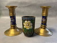 Frontal view of two brass candleholders and one cloisonné enamel bud vase showing floral designs and overall condition.