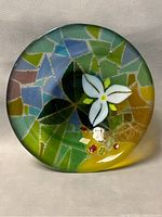 Top view of fused blown glass art bowl showing white flower with yellow center, green leaves and multicolored mosaic design.