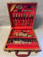 Opened wooden case showing entire 29-piece set of brass flatware with black handles and decorative brass tops, arranged inside red velvet lined sections including knives, forks, spoons and serving utensils.
