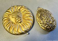 Front side of two items: gold-toned Estée Lauder Leo compact with lion face and rhinestones, and a smaller gold-toned ornate locket pendant with floral pattern.