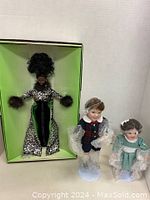 Barbie doll in box with two Ashton Drake porcelain dolls standing beside box on white surface