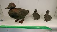 Cast iron duck figure and two rabbit figures displayed together on a white shelf with green painter's tape on edge.