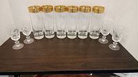 Front view of six tall clear beer glasses with thick gold decorative rims and four smaller etched flutes placed on a wood surface.