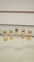 Photo showing five small glass vials with gold flakes sealed with cork or glass tops placed on a clear surface in front of a tiled wall.