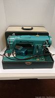 Front and base view of turquoise Manual Rotary portable sewing machine with foot pedal and cords, sitting in green base and beige carrying case behind.