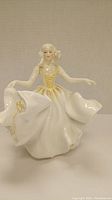 Front view of the smiling female figurine in a flowing white and yellow accented dress, arms outstretched.