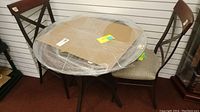 Photo of the round glass table with metal base wrapped in plastic along with two metal frame chairs with padded seats placed beside it.