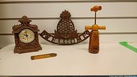 Four items displayed: ornate cast iron clock, cast iron welcome sign with pineapple design, wooden corkscrew, small wooden ruler/measuring tool.