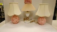 Three vintage table lamps on white background: two pink matching lamps with floral motifs and scalloped shades, and one white milk glass hobnail lamp with pleated shade and pink floral trim.
