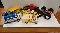 Photo showing all 7 vintage toy vehicles lined up, including a monster truck, school bus, news van, ambulance, tow truck, and the Ford Model A tow truck.
