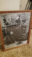 Framed black and white military photo showing two soldiers on a US Army vehicle with mounted machine gun. The photo is angled and resting against a wall with carpeted floor beneath it.