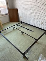 Metal bed frame positioned on carpeted floor next to wallpapered wall and window, showing full frame with wheels visible on corners and center.