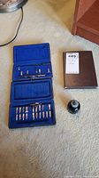 Blue velvet-lined case opened displaying multiple metal calligraphy nibs and two pen holders, along with bottle of ink and stamp album on floor.