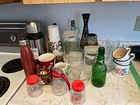 View showing assorted glassware, mugs, ceramic cups, vintage bottles, and vases arranged on a kitchen counter
