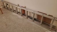 Full length view of aluminum extension ladder resting horizontally against a white wall on a concrete floor.
