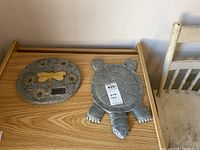 Two garden stone décor pieces on a wooden table, one oval with flowers and dragonfly, the other shaped like a turtle