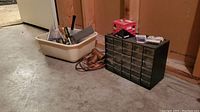View of the hardware organizer with drawers, white tub with tools, and copper coil on concrete floor.