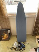 Ironing board with grey cover standing upright near window with two irons and blow dryer placed at its base.