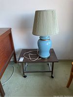 27 inch blue ceramic table lamp on small rolling wooden cart on carpeted floor next to wooden furniture.