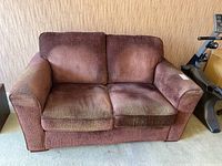 Front view of brown fabric two-seat love seat on light carpet against textured wallpaper. Shows seat cushion discoloration and wear.
