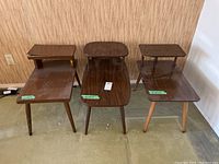 Shows three Mid-Century Modern end tables arranged side by side, two with dark tapered legs and one with lighter legs; tables exhibit wear and surface blemishes.