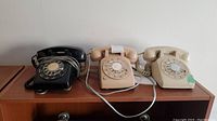 Three rotary dial telephones displayed on a wooden surface: black, cream, and beige in color.