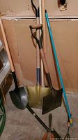 Photo of three shovels with wooden handles and different shaped blades leaning against a wall