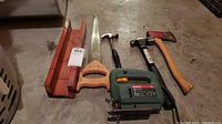 Full view of all tools including Bosch jig saw, saw, hammer, hatchet, and mitre box on concrete floor