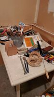 Photo showing a white folding table with an assortment of tools placed on it. Includes wrenches, pliers, coiled rope, chisels, paint brushes, metal bucket with miscellaneous tools, and boxes of supplies.
