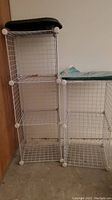 Two white wire cubby organizers side by side, each with three shelves, one has a black cushion on top, and a folded green tarp on the right organizer.