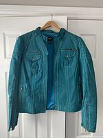 Full front view of blue suede leather women's jacket, hangs on wooden hanger against white door, showing front zipper and four pockets with zipper and snap button details.