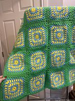 Front view of the crochet afghan blanket hanging, showing the square motifs with yellow, blue, and green yarn.